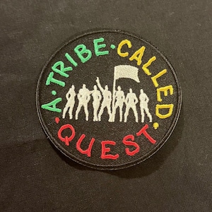 Puede incluir: Parche bordado circular con el texto "A Tribe Called Quest" en verde, amarillo y rojo. El parche presenta un grupo de figuras silueteadas y una bandera sobre un fondo negro.