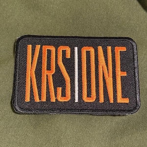 Puede incluir: Un parche rectangular con el texto "KRS ONE" en letras naranjas, separado por una línea blanca vertical. El parche está bordeado en negro y sobre un fondo verde oliva.