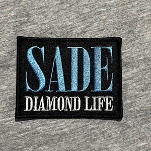 Op de afbeelding: Een zwart rechthoekig embleem met het woord "SADE" in lichtblauwe, gestileerde letters. Daaronder staan de woorden "DIAMOND LIFE" in witte hoofdletters. De patch staat op een grijze stoffen achtergrond.