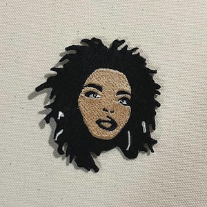 May include: Embroidered patch depicting a woman's face with black, curly hair. The face is tan with black outlines and blue eyes. The patch is set against a beige backdrop.