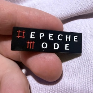 May include: A black rectangular pin with the text "DEPECHE MODE" in white letters. The pin has red accents, including a hashtag symbol and three vertical lines. The pin is held against a light purple fabric.