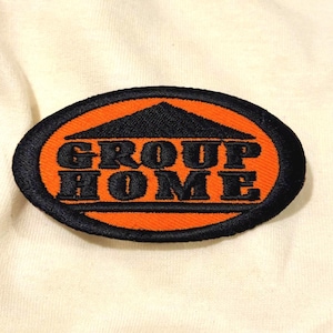 May include: An oval-shaped embroidered patch featuring a black border and an orange background. The patch displays a black triangular roof icon above the black text "GROUP HOME" and a horizontal black line at the bottom of the lettering.