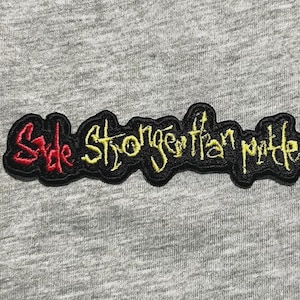 May include: Embroidered patch with the text "Style Stronger Than Mettle" in yellow and red on a black border. The patch is on a gray fabric background. The patch is approximately 4 inches long.