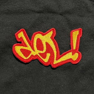 Op de afbeelding: Een rood en geel geborduurde patch met het woord "LOL" in een gestileerd lettertype. De patch heeft een rode rand en gele letters. De patch staat op een zwarte achtergrond.