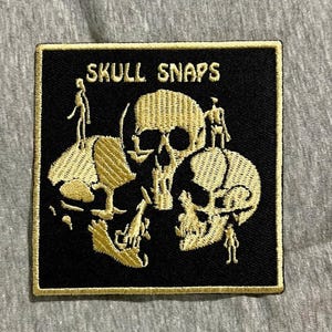 Op de afbeelding: Zwart vierkantige patch met goud geborduurde schedels en de tekst "SKULL SNAPS". De patch heeft drie schedels met kleine figuren erop. De patch heeft een gouden rand.