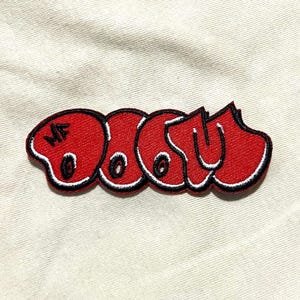 Może przedstawiać: Czerwona, haftowana naszywka z napisem "DOOM" w stylu graffiti. Naszywka ma czarny kontur z białą obwódką. Litery "MF" znajdują się w lewym górnym rogu naszywki.