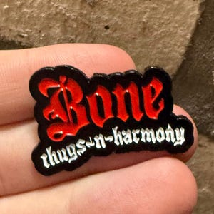 May include: A black and red enamel pin with the words "Bone thugs-n-harmony" in a gothic font. The pin has a black border and is held in a hand against a brick background.