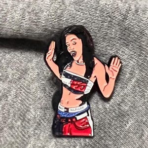 Pode incluir: Um alfinete colorido com uma ilustração de desenho animado de uma pessoa usando um top cropped e uma saia. A parte superior tem um design vermelho, branco e azul. A saia é vermelha e azul. O alfinete está em um fundo cinza.