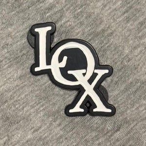 May include: Close-up of a gray fabric with a black and white "LOX" emblem. The emblem features the letters "L", "O", and "X" in a stylized design with a black outline and white fill.