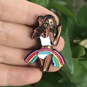 May include: A metal pin featuring a woman in a white top and rainbow skirt. The pin has a brown, gold, and rainbow color scheme. The woman is holding a red object to her mouth.