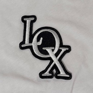 May include: A black and white embroidered patch with the letters "LOX". The letters are in a stylized font with a white outline and are sewn onto a black background. The patch is on a white fabric.