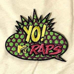 Puede incluir: Parche bordado con las palabras "YO! MTV RAPS" en amarillo y rojo, dentro de un bocadillo. El bocadillo tiene un fondo morado con lunares verdes y un contorno negro. También es visible el logotipo de MTV.