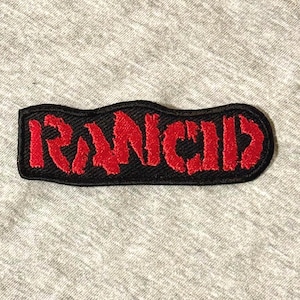 May include: A black embroidered patch with the word "RANCID" in red, stylized lettering. The patch has a black border and is set against a gray fabric background. The patch is rectangular with rounded edges.