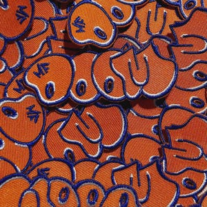 May include: A collection of orange embroidered patches featuring a smiling face design. Each patch has a white border and a blue outline. The patches are closely arranged, forming a textured pattern.