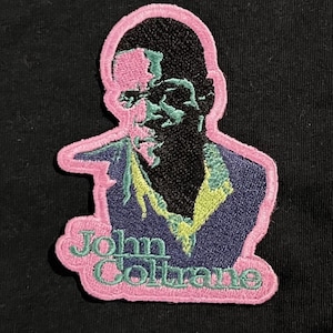 May include: Embroidered patch featuring a stylized portrait of John Coltrane. The patch has a pink border and includes the name "John Coltrane" in green script. The portrait uses black, blue, green, and pink colors.