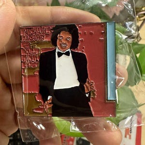 May include: A square pin featuring Michael Jackson in a black tuxedo and bow tie, with the text "MICHAEL JACKSON OFF THE WALL". The pin has a red and blue background.