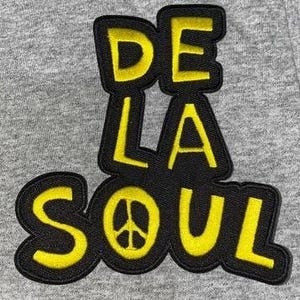 May include: Embroidered patch with the text "DE LA SOUL" in yellow letters outlined in black. The "O" in "SOUL" features a peace sign. The patch is on a gray fabric background.