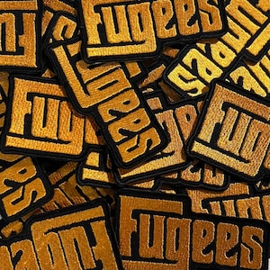 Può includere: Una collezione di toppe ricamate con la scritta "Fugees" in filo dorato su sfondo nero. Le toppe sono quadrate con angoli arrotondati e un bordo spesso. Le toppe sono sparse sulla superficie.