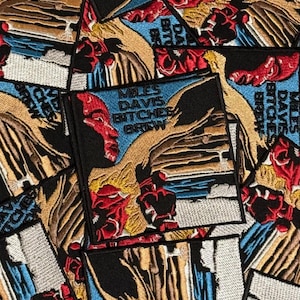May include: Embroidered patches featuring the album art for Miles Davis's "Bitches Brew." The patches display a portrait of a person with red and black accents, set against a blue background. The text "Miles Davis Bitches Brew" is also visible.