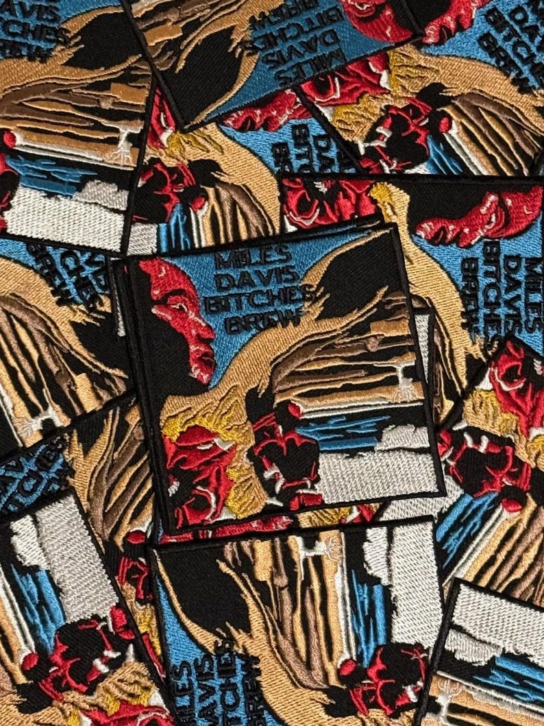 May include: Embroidered patches featuring the album art for Miles Davis's "Bitches Brew." The patches display a portrait of a person with red and black accents, set against a blue background. The text "Miles Davis Bitches Brew" is also visible.