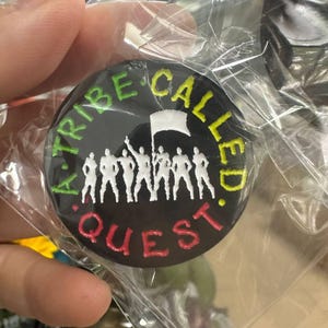 Puede incluir: Un botón redondo negro con el texto "A Tribe Called Quest" en verde, amarillo y rojo. El botón presenta una silueta blanca de personas sosteniendo una bandera. El botón está en un envoltorio de plástico transparente.
