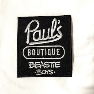 May include: A black square patch with white embroidered text. The text reads "Paul's BOUTIQUE" in a stylized font, with "BEASTIE BOYS" below. The patch is set against a white background.