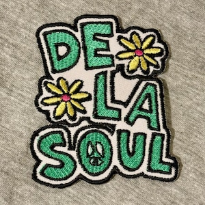 Op de afbeelding: Geborduurde patch met de tekst "DE LA SOUL" in groen, omlijnd met zwart, op een witte achtergrond. Twee gele bloemen met roze centra en een vredesteken zijn in het ontwerp opgenomen. De patch ligt op een grijze stof.
