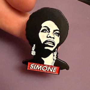 Puede incluir: Un pin de esmalte en blanco y negro con un retrato de Nina Simone con afro y pendientes. El pin tiene una pancarta roja con el nombre "SIMONE". El pin está sujeto por los dedos de una persona. El pin es un retrato de la cantante.