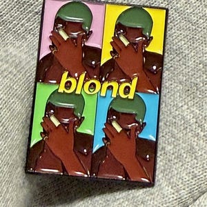 May include: A rectangular pin featuring a four-panel design with a portrait of a person with green hair and brown skin. The word "blond" is written in yellow. Each panel has a different colored background: pink, yellow, green, and blue.