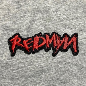 Puede incluir: Un parche bordado rojo con la palabra "REDMAN" en una fuente estilizada y dentada. El parche tiene un borde negro y está sobre un fondo de tela gris. El texto parece estar cosido.