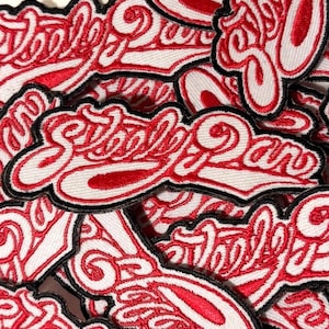 May include: Embroidered patches with the text "School Daze" in a cursive font. The patches feature a white background with red lettering and a red border, outlined in black. The patches are piled together.