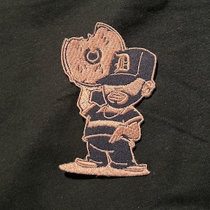 May include: Embroidered patch featuring a cartoon character in a striped shirt and a baseball cap with a "D" on it. The character is holding a donut with a bite taken out of it. The patch has a brown and navy blue color scheme.