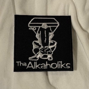 Könnte beinhalten: Schwarzer quadratischer Aufnäher mit einem weißen, gestickten Design einer Person auf einer Toilette, die ein Mikrofon hält. Der Text "Tha Alkaholiks" befindet sich darunter.