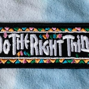 Könnte beinhalten: Schwarzer rechteckiger Aufnäher mit dem weißen gestickten Schriftzug "Do The Right Thing". Der Aufnäher ist mit einem farbenfrohen Design aus Dreiecken und floralen Akzenten in Orange, Rosa und Türkis eingefasst.