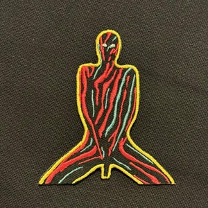 Könnte beinhalten: Gestickter Aufnäher mit einer stilisierten knienden Figur. Die Figur hat ein rot-schwarzes Streifenmuster mit blauen Akzenten, umrandet von Gelb. Der Aufnäher befindet sich auf einem schwarzen Hintergrund.