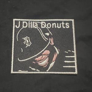 May include: Embroidered patch featuring the text "J Dilla Donuts" above a stylized image of a baseball cap with the Detroit Tigers logo. The patch is black with white, pink, and gray details and a white border.