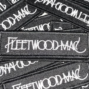 May include: Black rectangular embroidered patches with the text "FLEETWOOD MAC" in white cursive lettering. The patches are stacked on top of each other, with the top patch in focus. The patches are likely for clothing or accessories.