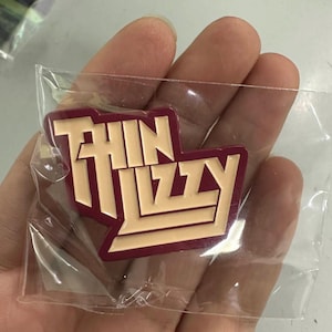 Puede incluir: Un pin con el logotipo de Thin Lizzy en burdeos y beige, dentro de un envoltorio de plástico transparente. El pin presenta el nombre de la banda en una fuente estilizada. El pin está en la mano de una persona.