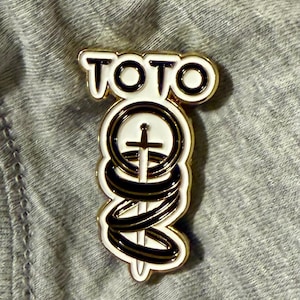 May include: A gold-coloured pin featuring the word "TOTO" in white with a black outline. The pin depicts a sword through a circle, encircled by black and gold swirls, set against a white backdrop.