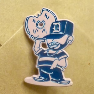 May include: A blue and white enamel pin featuring a cartoon character holding a donut. The character wears a baseball cap with a "D" on it, a striped shirt, and a chain necklace. The pin has a pink border.