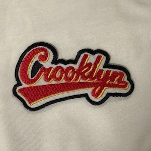 May include: An embroidered fabric patch featuring the word Crooklyn in red cursive script with a gold outline and a thick black border. The text is underlined by a bold red swoosh with a gold edge, all set against a plain off-white background.