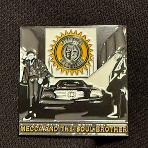 May include: Square pin featuring a black car, two figures, and a logo with a face. The pin has a black, white, and gold color scheme. The text "MECCA AND THE SOUL BROTHER" is at the bottom.