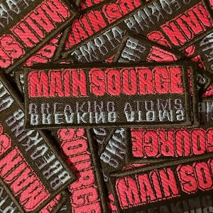 Puede incluir: Parches bordados rectangulares negros con el texto "MAIN SOURCE" en rojo. Debajo, la frase "BREAKING ATOMS" está bordada en azul claro. La imagen muestra una colección de estos parches.