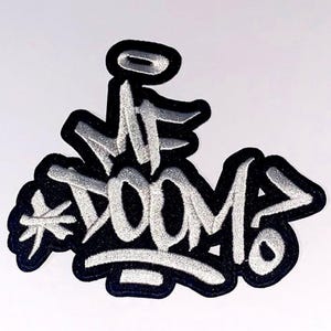 Peut inclure: Écusson brodé avec le texte "MF DOOM" dans un style graffiti. L'écusson présente des lettres blanches avec un contour noir et un fond noir. L'écusson a une bordure blanche et un motif d'étoile.