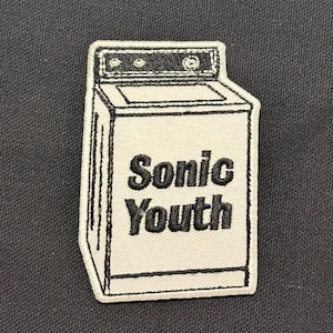 Puede incluir: Una taza de cerámica blanca con un asa negra y el texto "But First, Coffee" en negrita negra. La taza se encuentra sobre un fondo borroso, sugiriendo un ambiente acogedor.