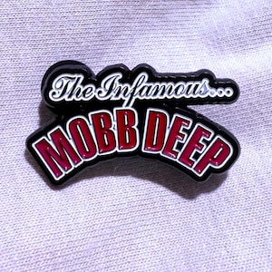 May include: A black and red enamel pin featuring the text "The Infamous..." above "Mobb Deep" in a stylized font. The pin has a white border and is set against a light gray fabric background.