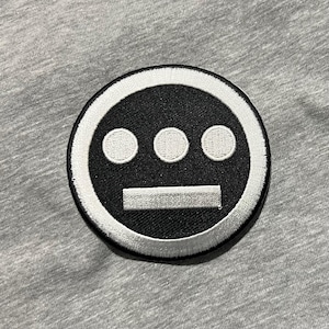 May include: A circular black and white embroidered patch featuring a stylized face design. The design includes three white dots above a horizontal white bar, all set against a black background. The patch is on a gray fabric.
