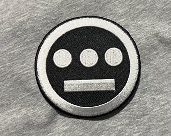 May include: A circular black and white embroidered patch with a stylized face design. The design features three white dots above a horizontal white bar on a black background. The patch is set against a grey fabric.