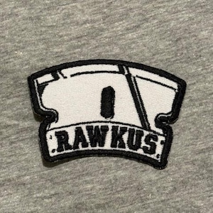May include: A white and black embroidered patch with the text "RAWKUS" in a stylized font. The patch has a black border and is set against a gray fabric background.