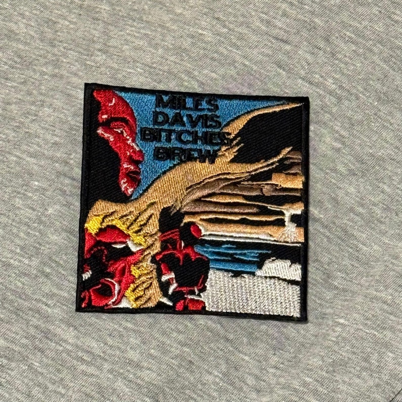 May include: Embroidered patch featuring a portrait of Miles Davis with the text "Miles Davis Bitches Brew." The patch has a black border, a blue sky, and a landscape design with red, yellow, and brown accents.